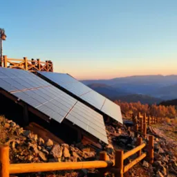 Dwa panele słoneczne zainstalowane na zboczu wzgórza, z widokiem na zalesione góry o zachodzie słońca. W tle drewniana balustrada i sylwetki ludzi.