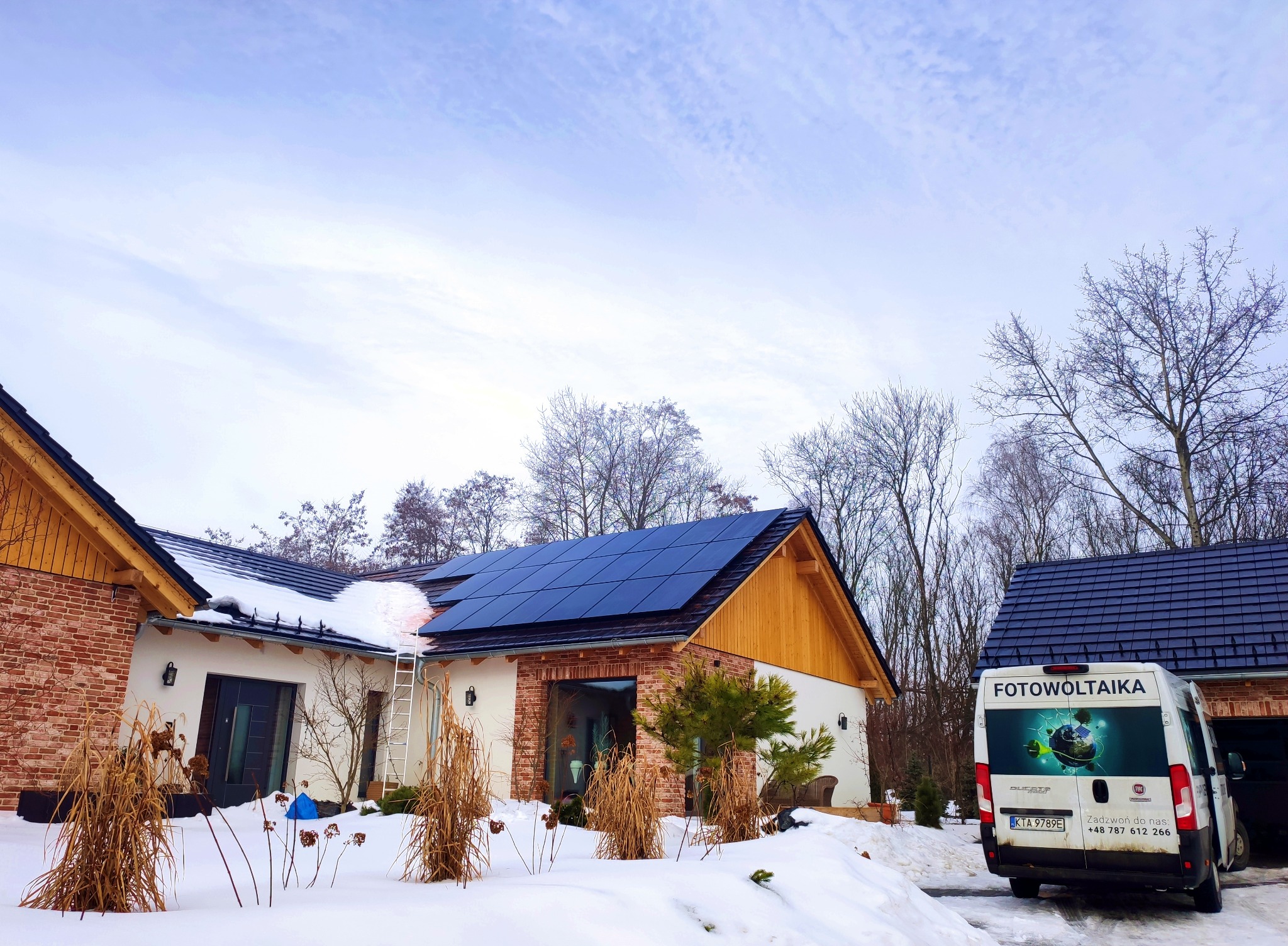 Dom jednorodzinny z panelami słonecznymi na dachu, częściowo pokrytym śniegiem, oraz zaparkowanym obok samochodem dostawczym z napisem na karoserii. Na dachu stoi drabina.