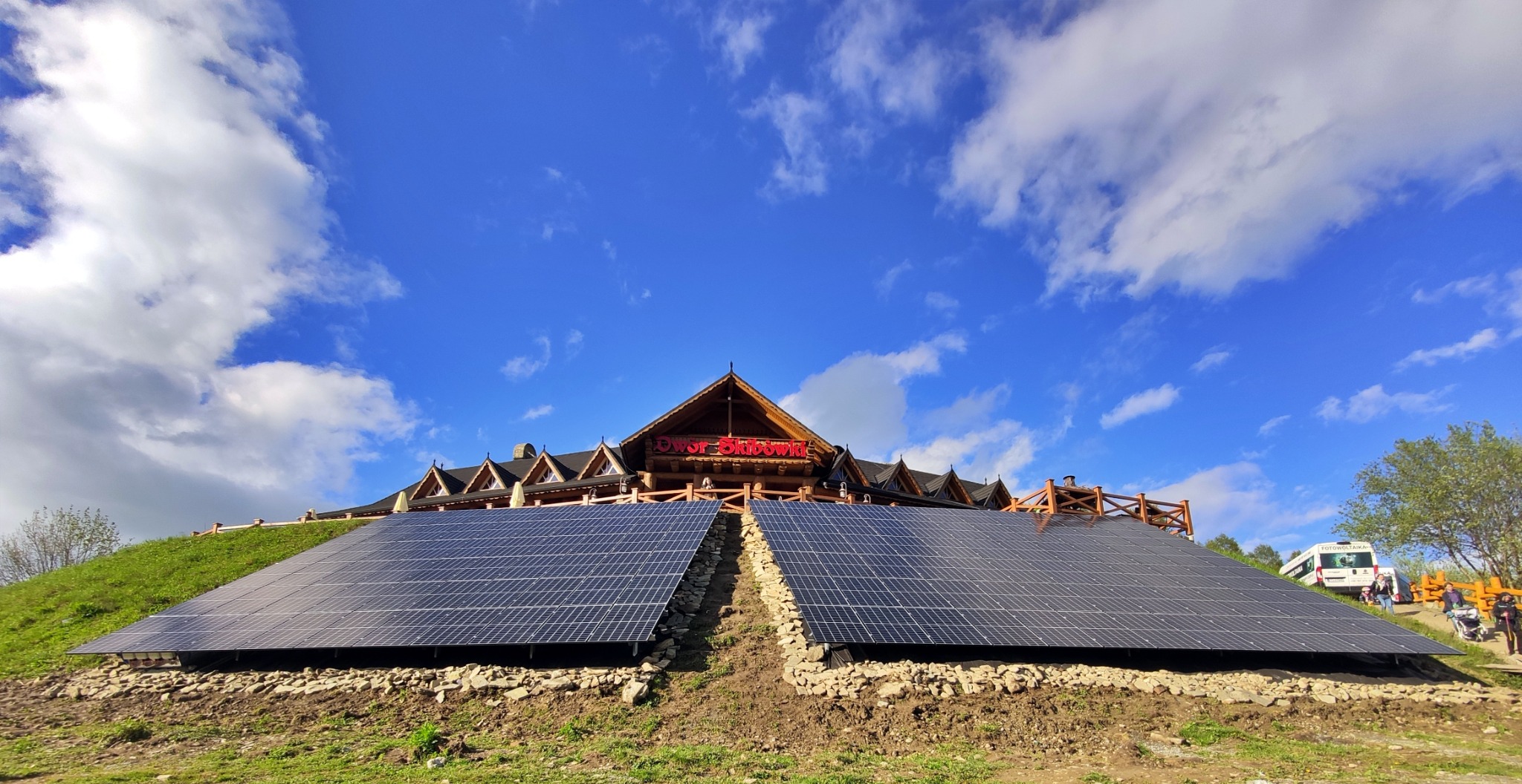Panele słoneczne zamontowane na zboczu wzgórza przed drewnianym budynkiem z napisem 'Dwór Skibówki' na tle błękitnego nieba z chmurami.
