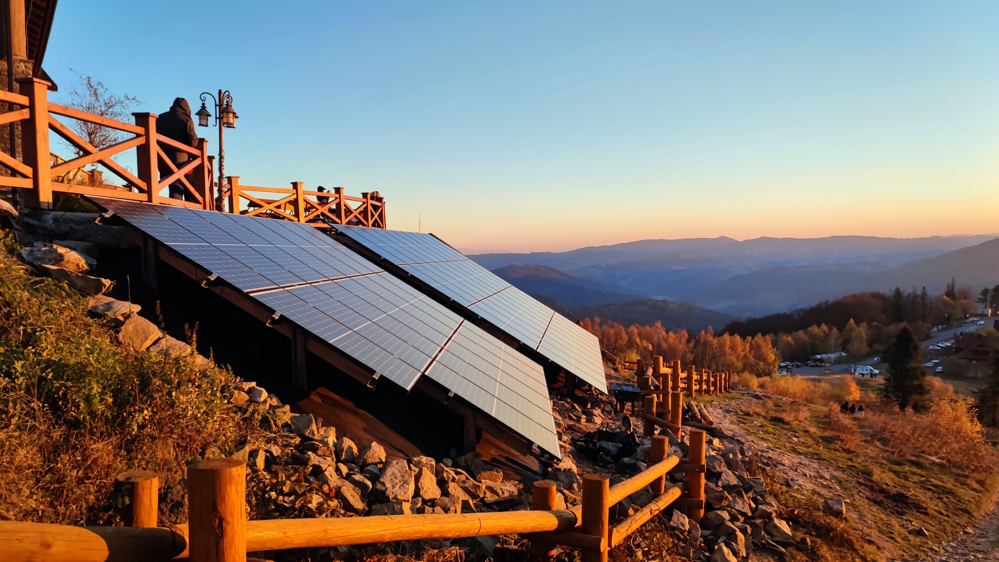 Dwa panele słoneczne zainstalowane na zboczu wzgórza, z widokiem na zalesione góry o zachodzie słońca. W tle drewniana balustrada i sylwetki ludzi.