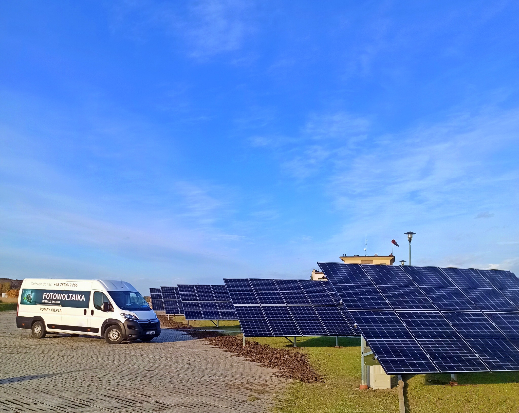 Rząd paneli fotowoltaicznych na trawie, z białym vanem z napisem 'Fotowoltaika' i numerem telefonu w tle, na tle błękitnego nieba z chmurami.