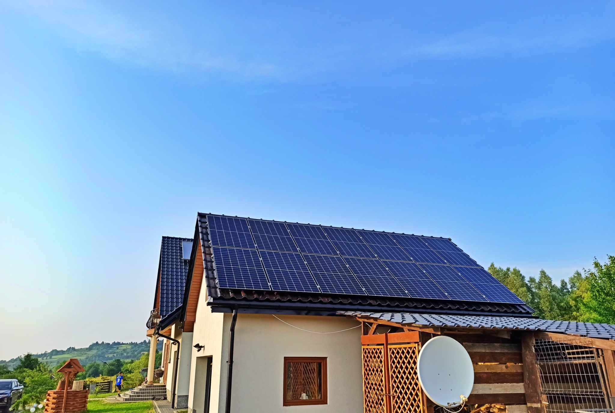 Dom jednorodzinny z panelami słonecznymi na dachu, widok z dołu na tle błękitnego nieba, w tle wzgórza i las.