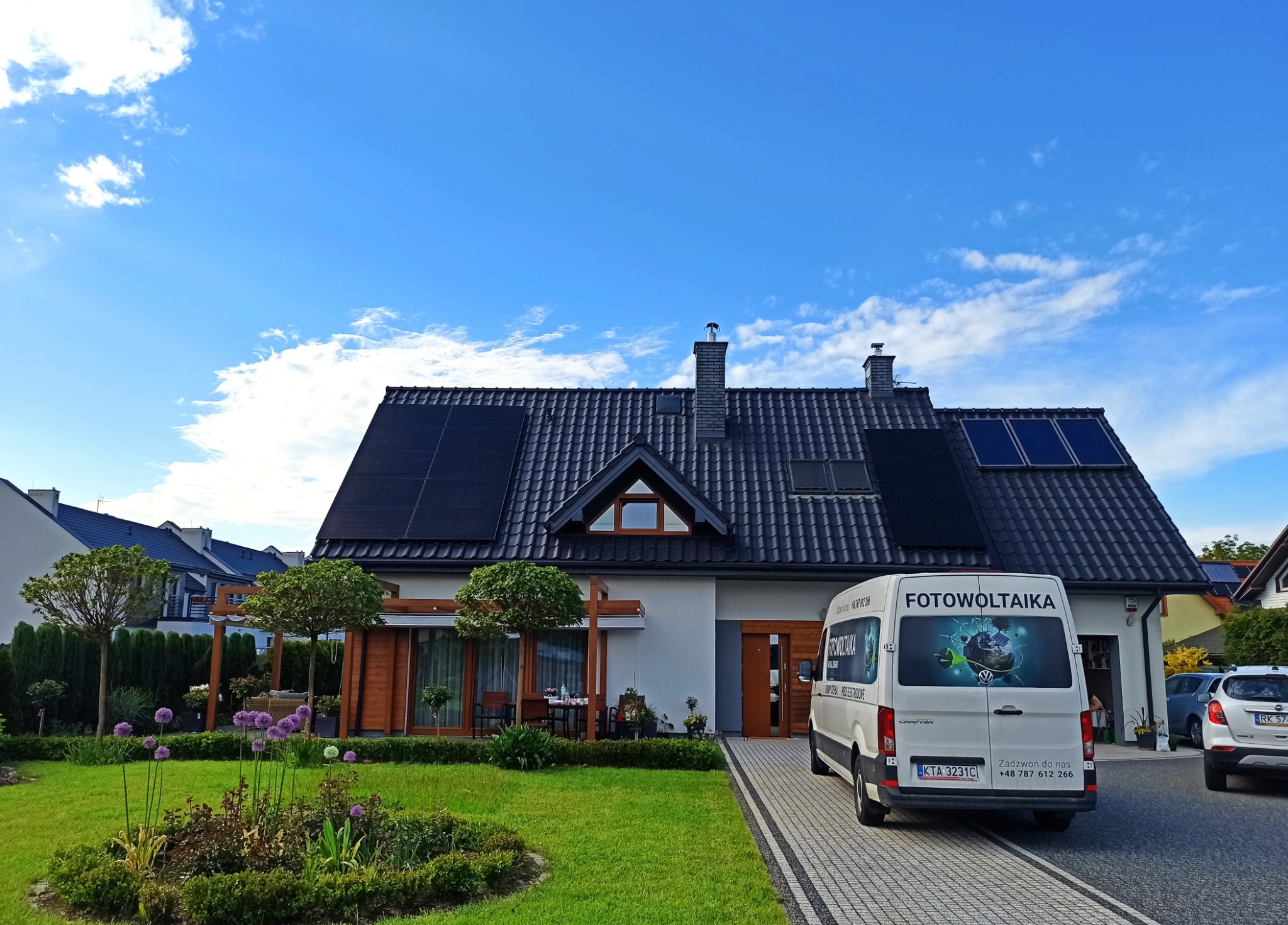 Dom jednorodzinny z panelami słonecznymi na dachu, zaparkowany biały bus z reklamą firmy, zadbany trawnik z kwiatami na pierwszym planie, błękitne niebo.