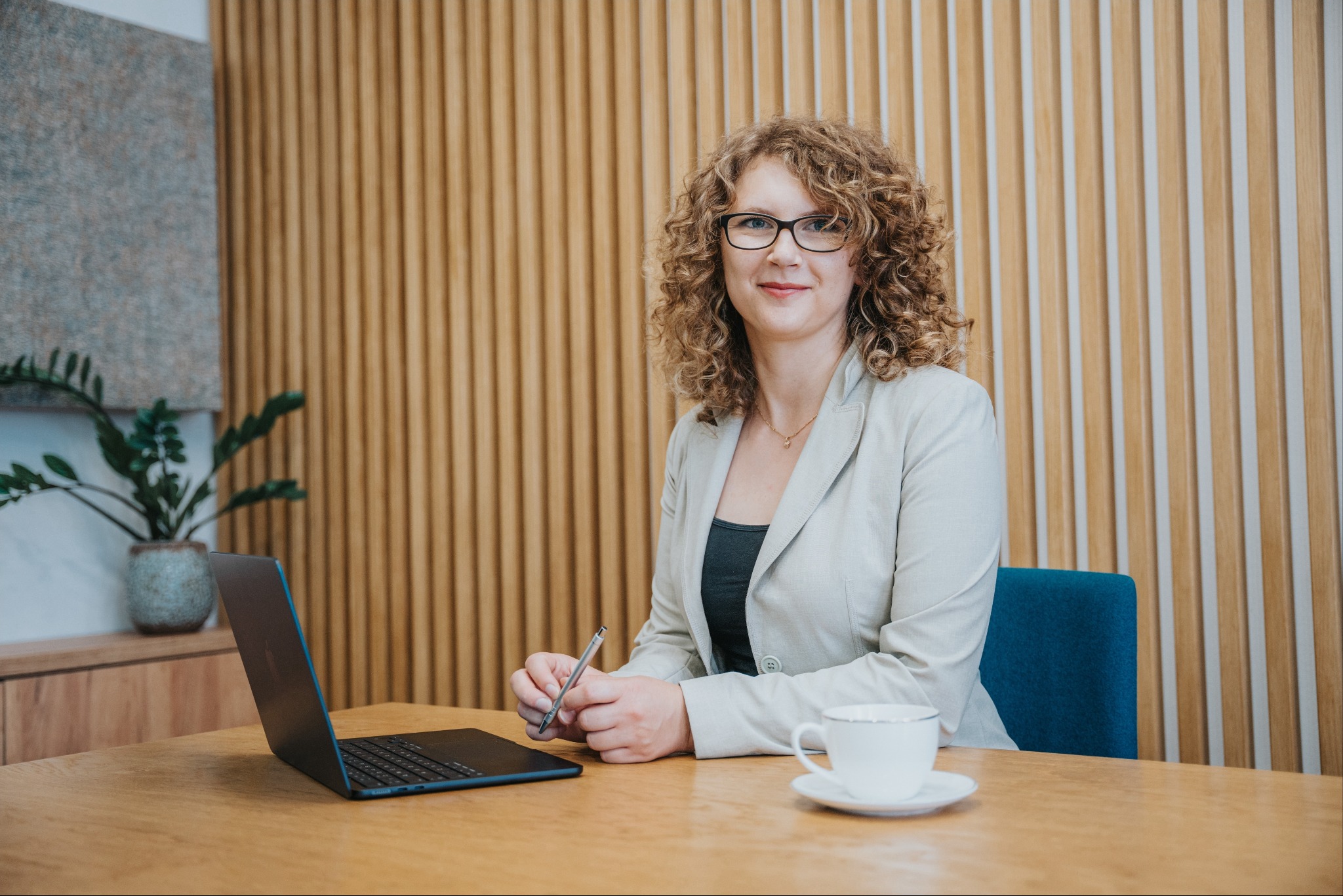 Kobieta w okularach, siedząca przy biurku z laptopem i filiżanką, trzymająca długopis w dłoniach, z drewnianą ścianą i rośliną w tle.