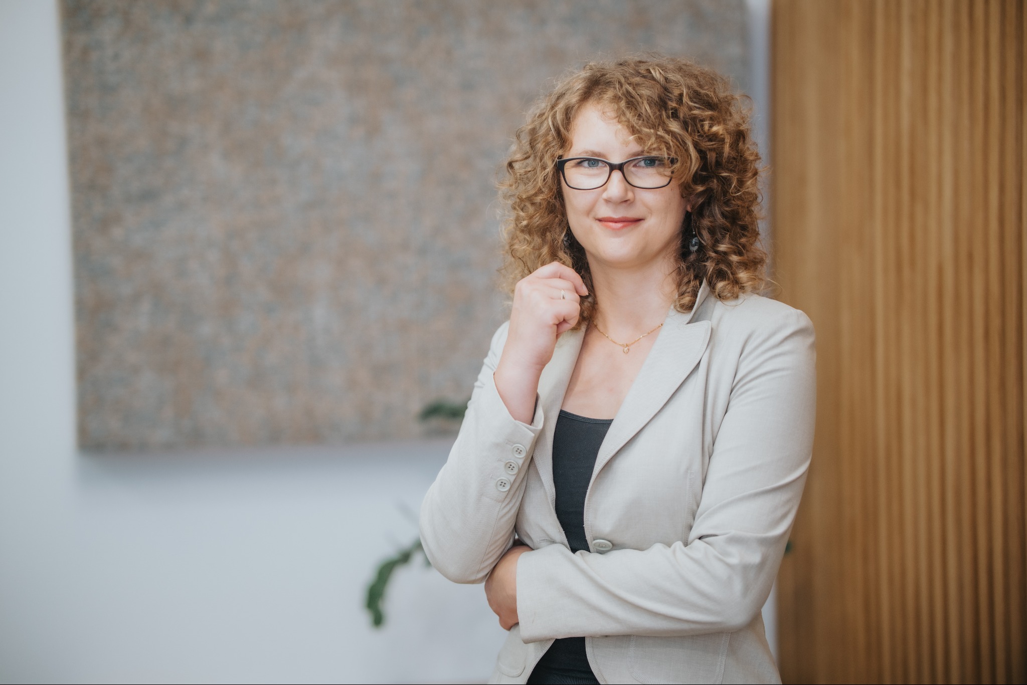 Portret kobiety w okularach i beżowym żakiecie, z ręką pod brodą, na neutralnym tle z elementami dekoracyjnymi.