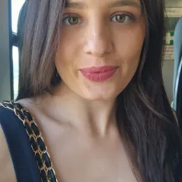 Portret uśmiechniętej kobiety z ciemnymi włosami i złotym łańcuszkiem torebki na ramieniu, zrobiony wewnątrz wagonu pociągu.