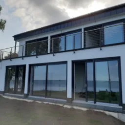 Nowoczesny dom z dużymi przesuwnymi oknami z czarnymi ramami i balkonem z metalową balustradą, widok na jezioro, drabina oparta o ścianę, trawnik w trakcie zakładania.