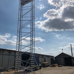 Lockhard Sp. z o.o. - Wysoki, metalowy podnośnik koszowy na kółkach z pracownikiem na platformie, na tle budynków gospodarczych i błękitnego nieba z chmurami.