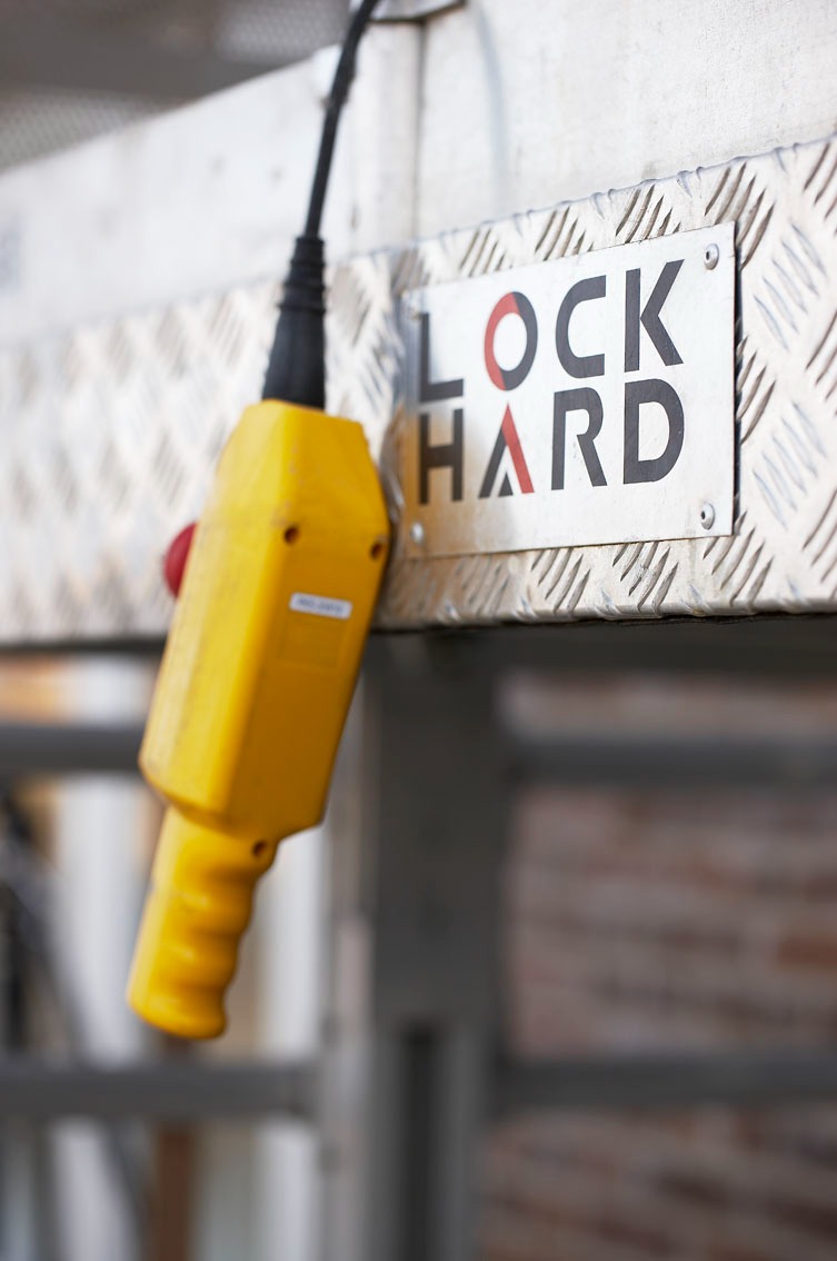 Żółty pilot sterowania z czerwonym przyciskiem, podłączony kablem, obok metalowej tabliczki z napisem 'LOCK HARD' na tle metalowej konstrukcji z kratownicą.