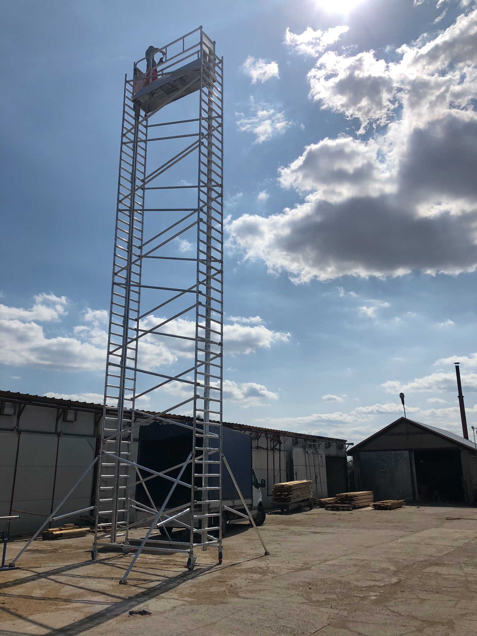 Wysoki, metalowy podnośnik koszowy na kółkach z pracownikiem na platformie, na tle budynków gospodarczych i błękitnego nieba z chmurami.