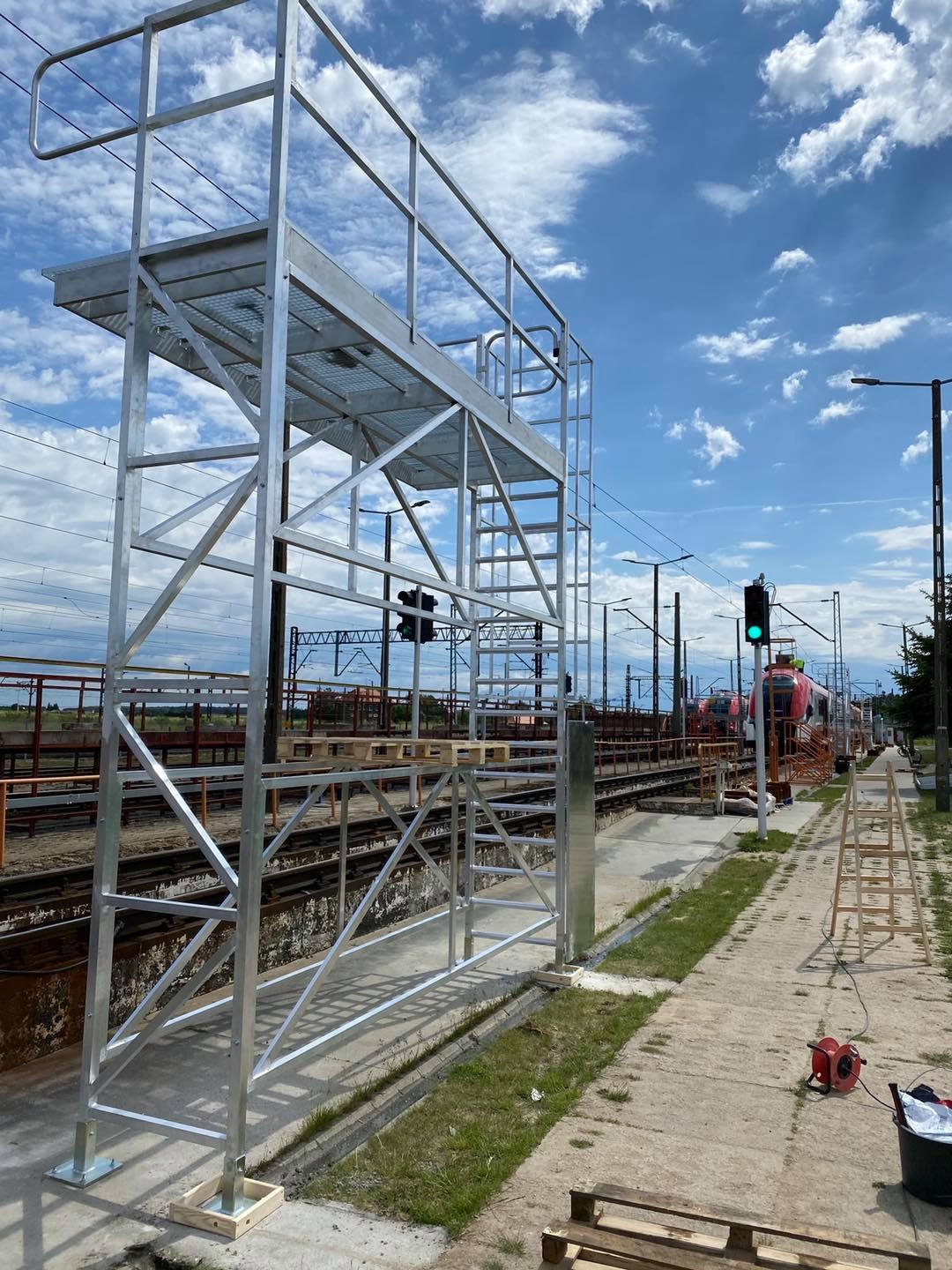 Metalowa konstrukcja platformy roboczej na stacji kolejowej, widoczny pociąg w tle, zielone światło semafora.