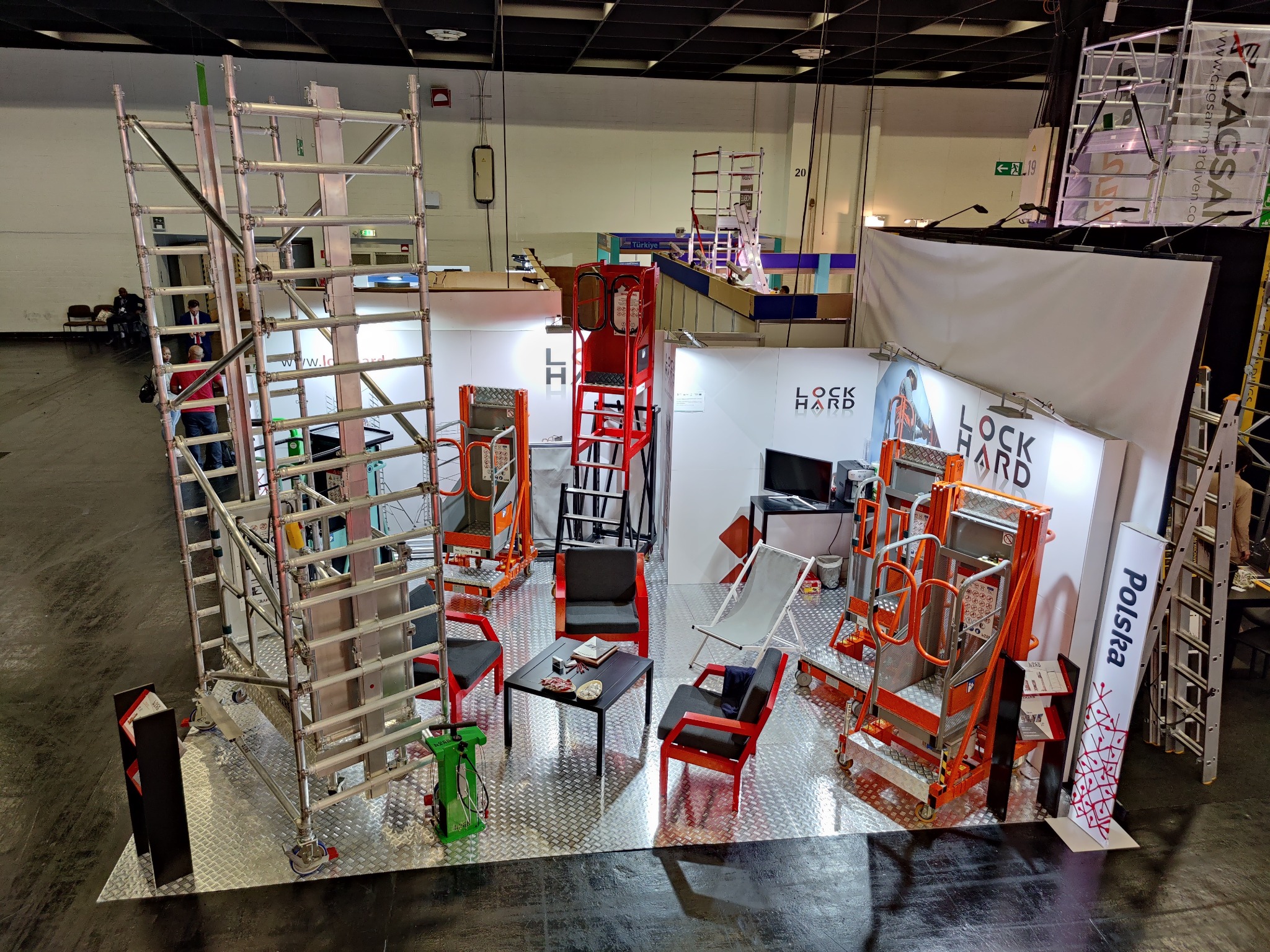 Ekspozycja na targach z różnymi modelami podestów roboczych, schodów i rusztowań w kolorach srebrnym, pomarańczowym i czerwonym, ustawionych na metalowej platformie. W tle widoczne stanowisko...