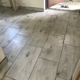 Ułożone podłużne płytki podłogowe imitujące drewno w odcieniach szarości, widoczne fragmenty ścian i futryny drzwi, buty robocze w przejściu.