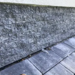 Szara elewacja z kamiennych płytek ułożonych poziomo, widoczne szare płyty chodnikowe i fragment białego tynku powyżej elewacji.