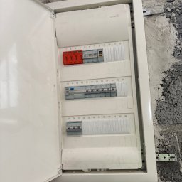E-TEAM Michał Potaczek - Otwarta, biała skrzynka elektryczna Hager z bezpiecznikami i numeracją 1-36, zamontowana na surowej, szarej ścianie. Widoczne elementy instalacji i mocowania.