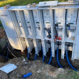 E-TEAM Michał Potaczek - Rozdzielnica elektryczna z podłączonymi niebieskimi rurami w ziemi, widoczne narzędzia i skrzynki. Prace przyłączeniowe w terenie otwartym.