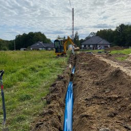 E-TEAM Michał Potaczek - Wykopany rów z niebieską rurą w ziemi, przygotowany pod instalację elektryczną. W tle koparka i słup wysokiego napięcia na tle pochmurnego nieba.