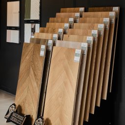 Pixel Dariusz Soboń - Ekspozycja paneli podłogowych w jodełkę, różne odcienie drewna, eleganckie stojaki z logo producenta, plakaty dekoracyjne na ścianie. Prezentacja oferty podłóg.