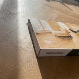 Pixel Dariusz Soboń - Ułożona podłoga w jodełkę, częściowo zakryta kartonem z wizualizacją wnętrza i napisem 'GLUE DOWN MULTICONTRACT'. W tle widoczna ściana w trakcie wykończenia.