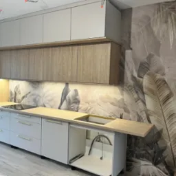 Nowoczesna kuchnia z szarymi szafkami, drewnianym blatem i tapetą w liście. Widoczne detale wykończenia i oświetlenie podszafkowe. Elementy dekoracyjne z drewna.