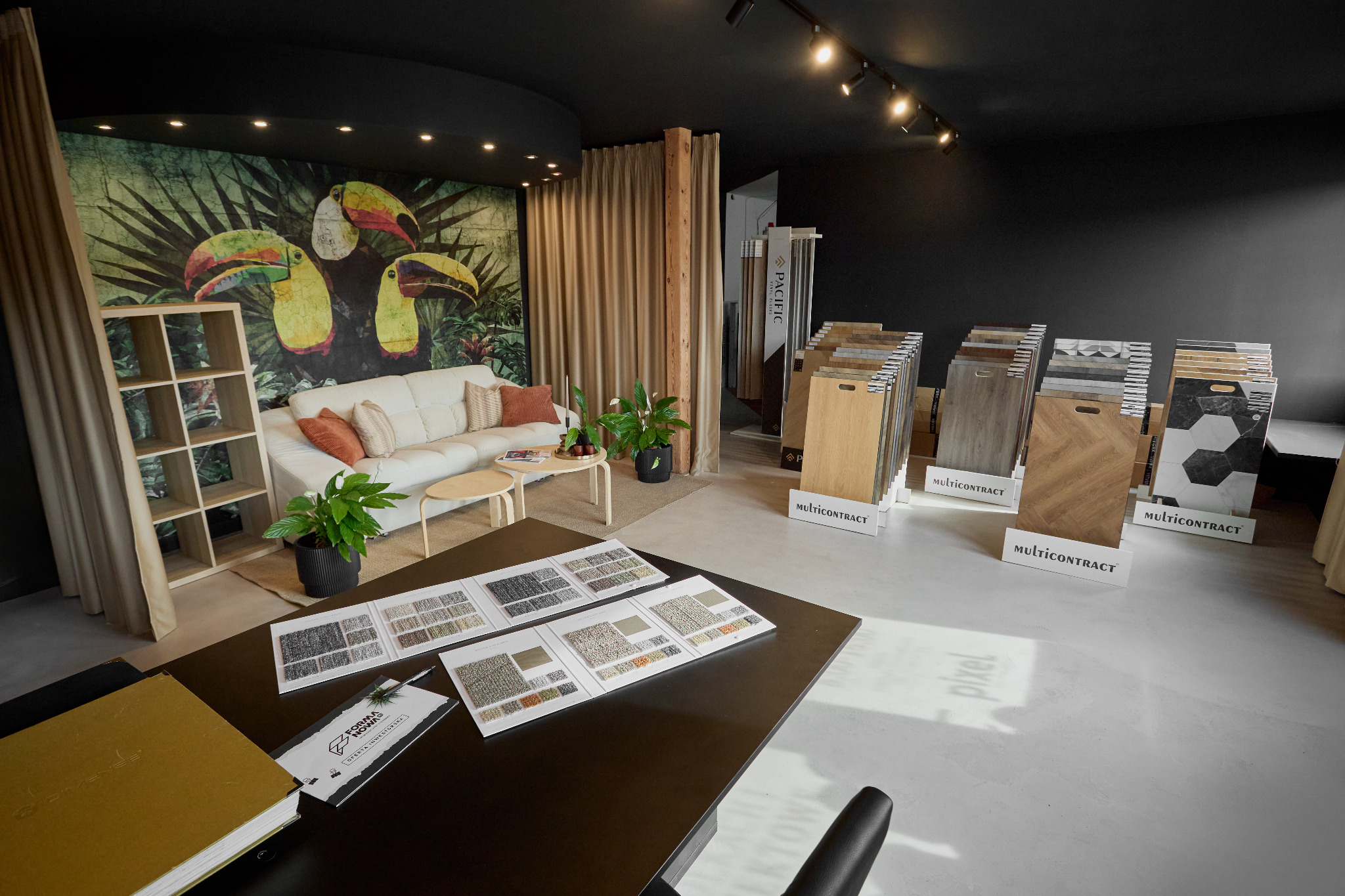 Salon z ekspozycją paneli podłogowych i wykładzin w Kołobrzegu. Widoczne wzorniki, próbki materiałów, sofa i tapeta z motywem tropikalnym.