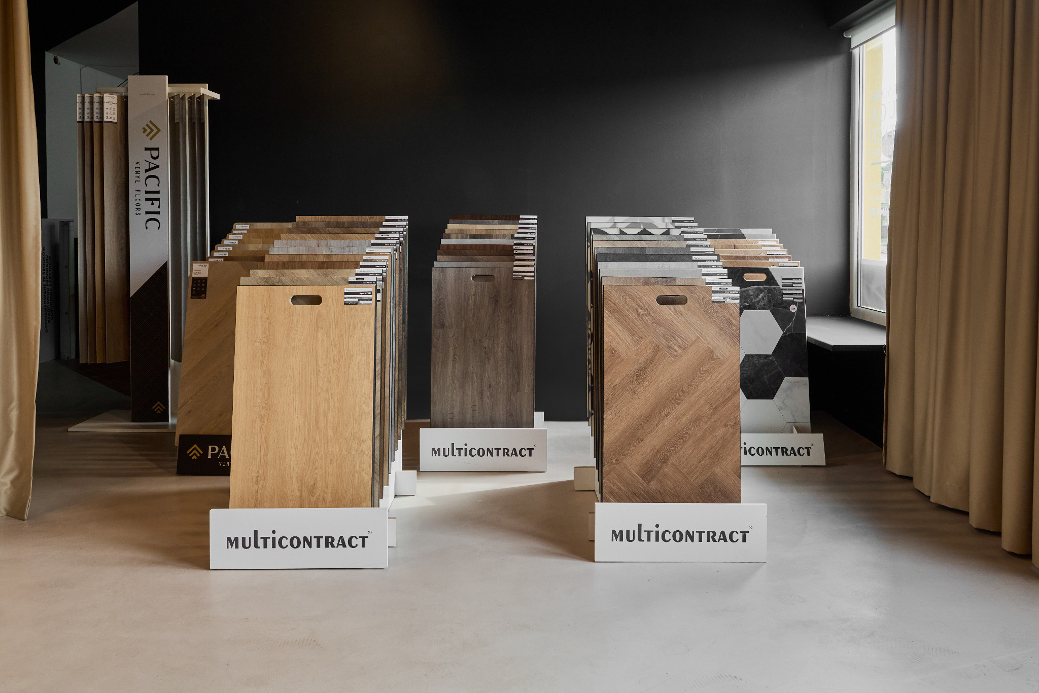 Ekspozycja różnorodnych paneli podłogowych w salonie: od jasnych dębowych po ciemne, wzorzyste. Widoczne logo producenta 'Multicontract' i stojak 'Pacific Vinyl Floors'.