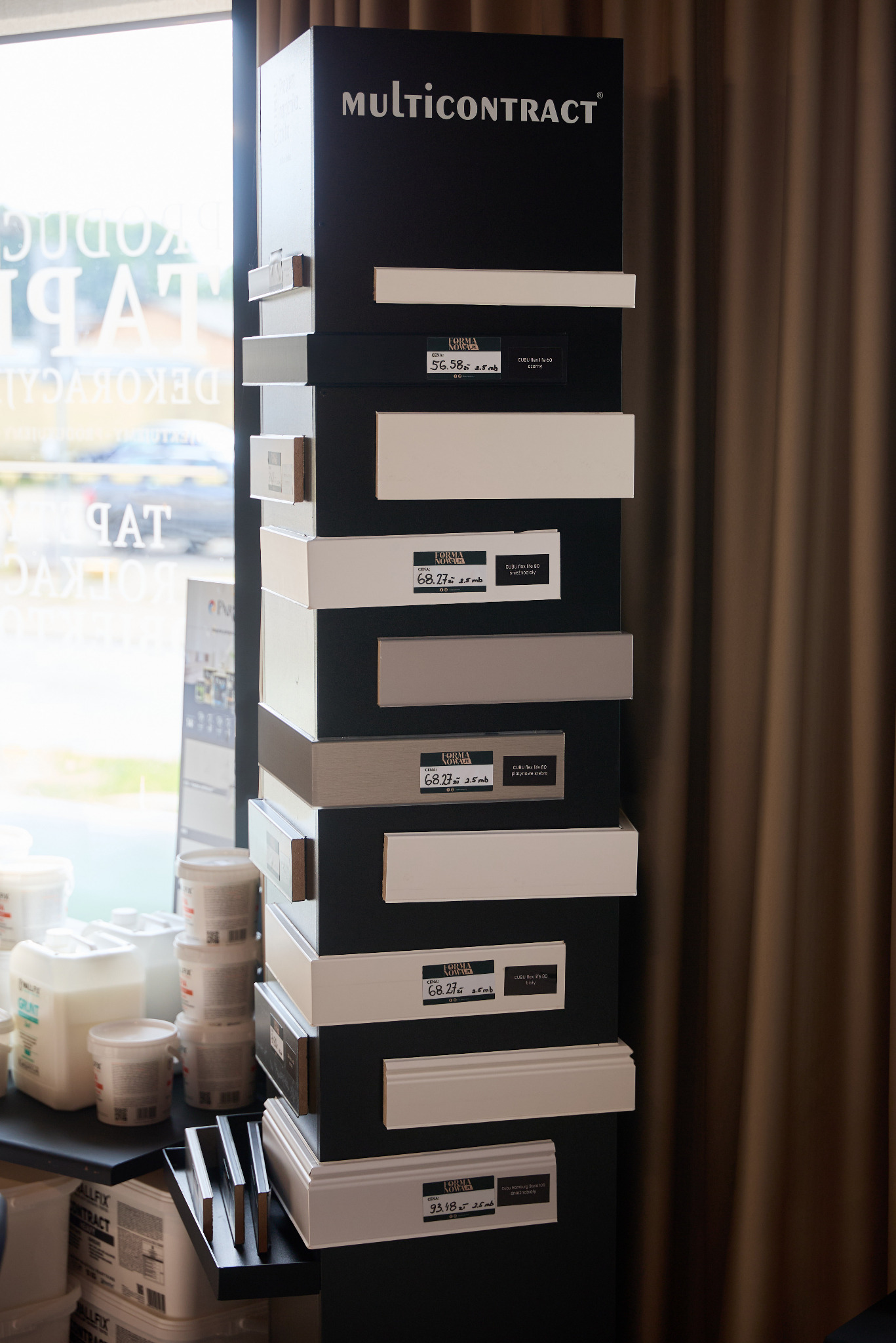 Ekspozycja próbek listew przypodłogowych w sklepie, z widocznymi cenami i nazwami modeli na czarnym stojaku. W tle okno i zasłony.