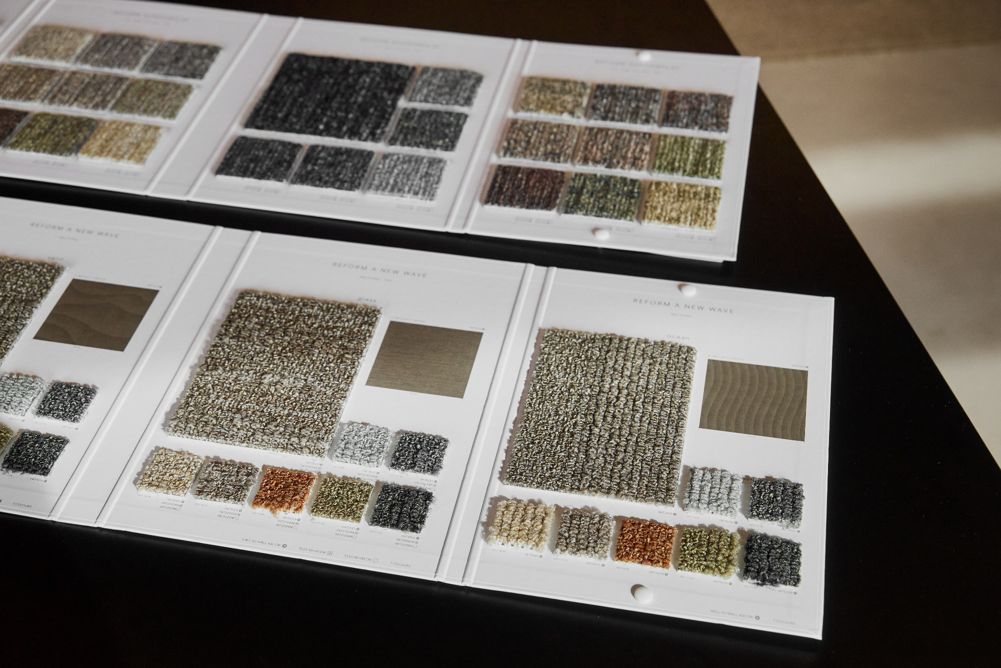 Katalog próbek wykładzin dywanowych 'Reform A New Wave' na czarnym stole. Widoczne różne kolory i tekstury. Prezentacja kolekcji.