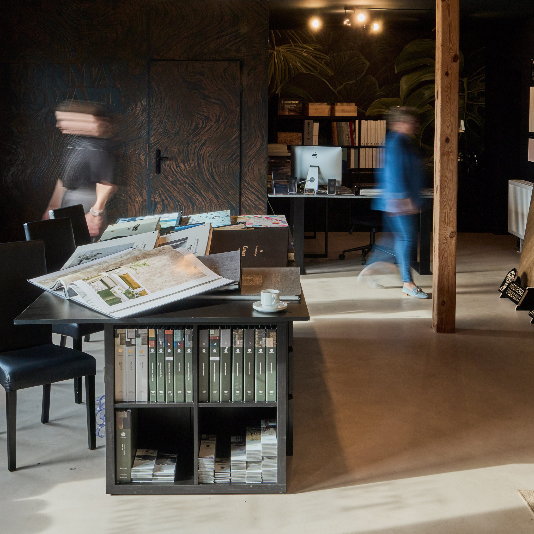 Biuro projektowe z katalogami, próbkami materiałów, biurkiem i komputerem. Osoby w ruchu, wnętrze z tapetą w tropikalne liście, ciepłe oświetlenie.