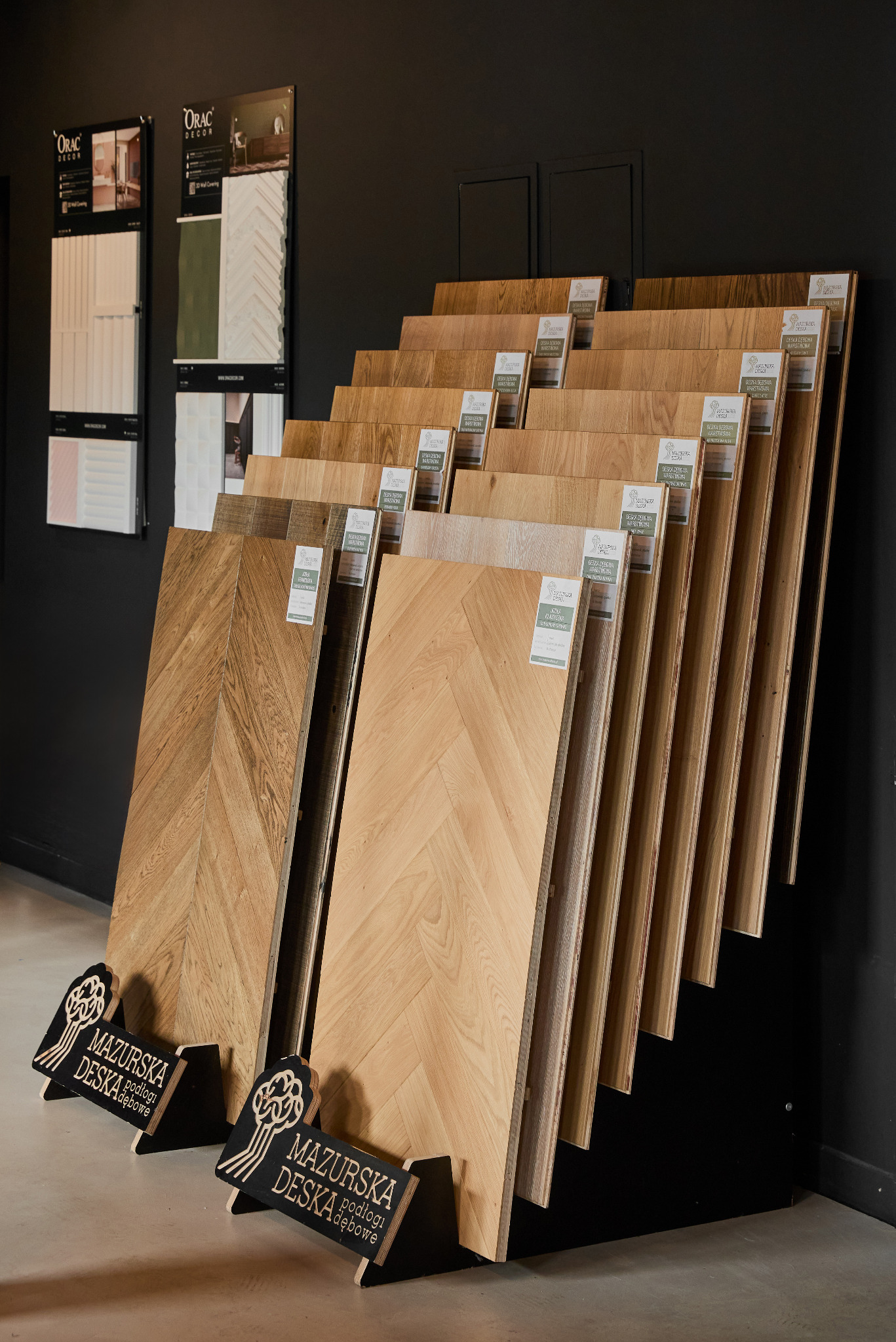 Ekspozycja paneli podłogowych w jodełkę, różne odcienie drewna, eleganckie stojaki z logo producenta, plakaty dekoracyjne na ścianie. Prezentacja oferty podłóg.