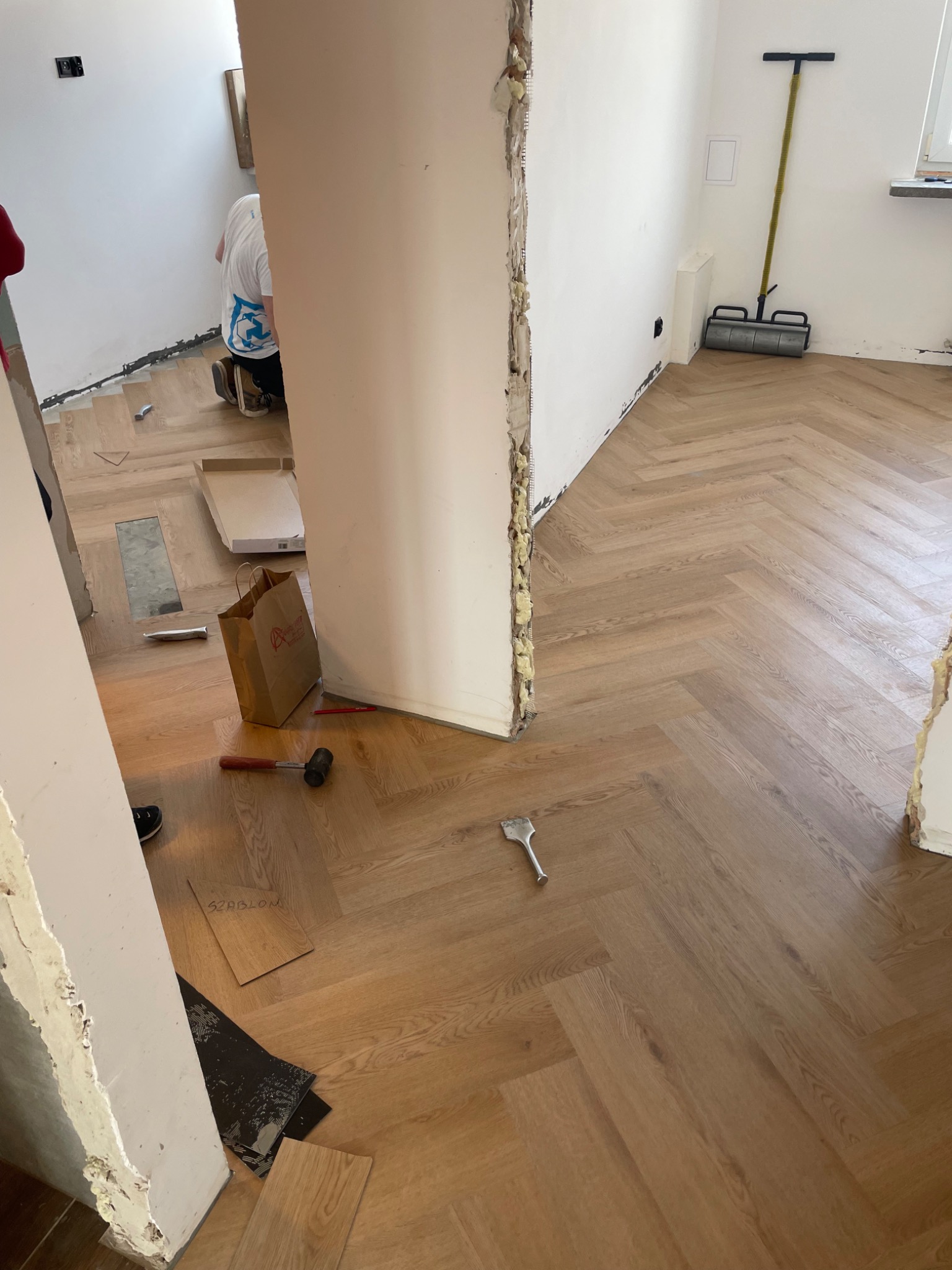 Układanie parkietu w jodełkę, widoczne narzędzia i materiały. Częściowo wykończona podłoga z jasnego drewna. W tle pracownik w trakcie montażu.