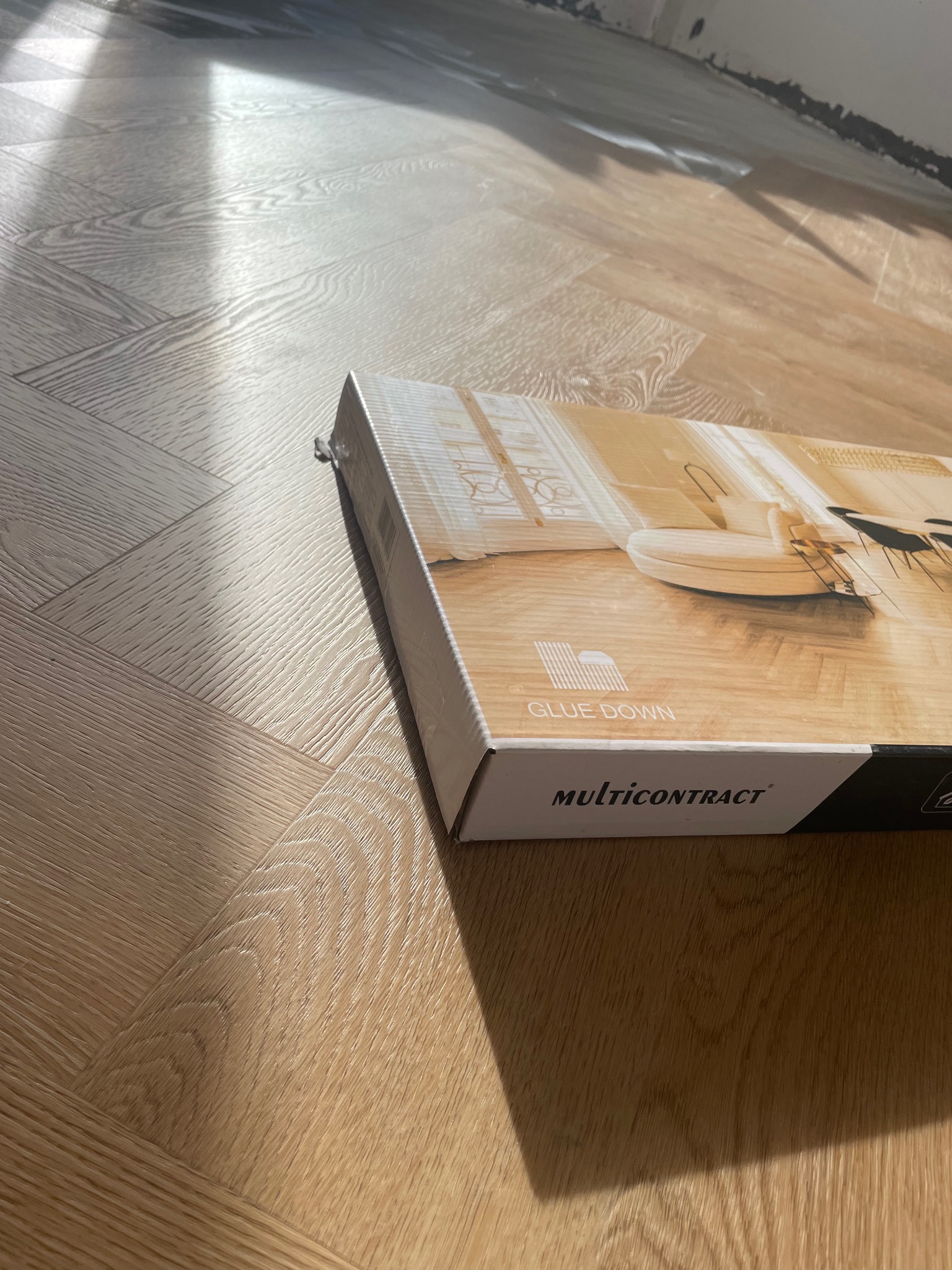 Ułożona podłoga w jodełkę, częściowo zakryta kartonem z wizualizacją wnętrza i napisem 'GLUE DOWN MULTICONTRACT'. W tle widoczna ściana w trakcie wykończenia.