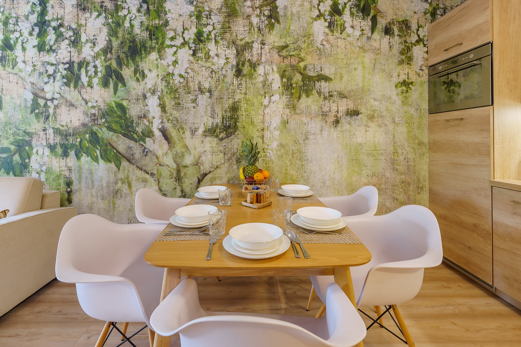 Stylowa jadalnia z drewnianym stołem, białymi krzesłami i tapetą z motywem drzewa. Oświetlenie punktowe podkreśla detale aranżacji.