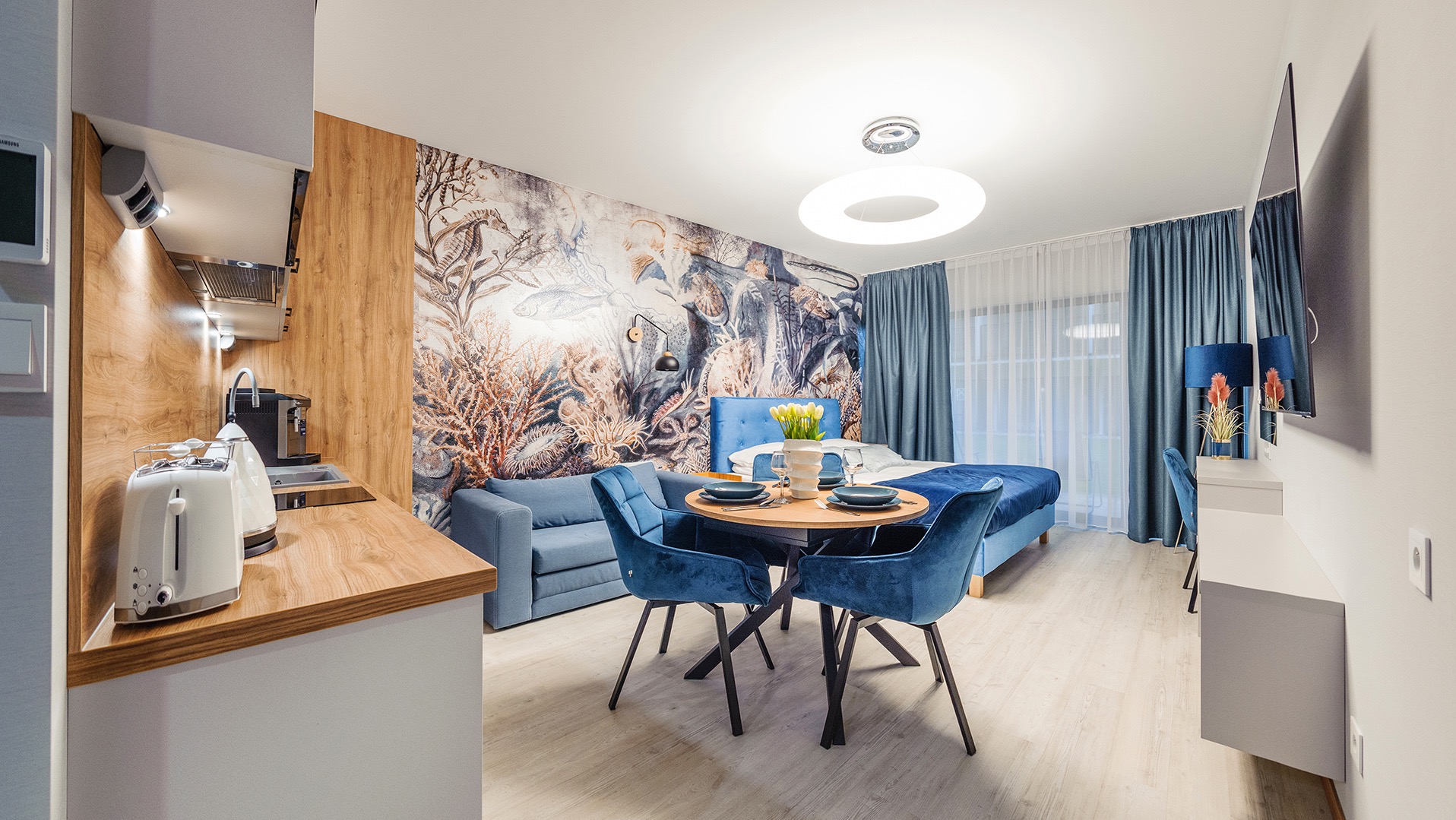 Nowoczesne wnętrze apartamentu z aneksem kuchennym, stołem jadalnym i łóżkiem, utrzymane w błękitnej kolorystyce, z tapetą w motywy morskie na ścianie.
