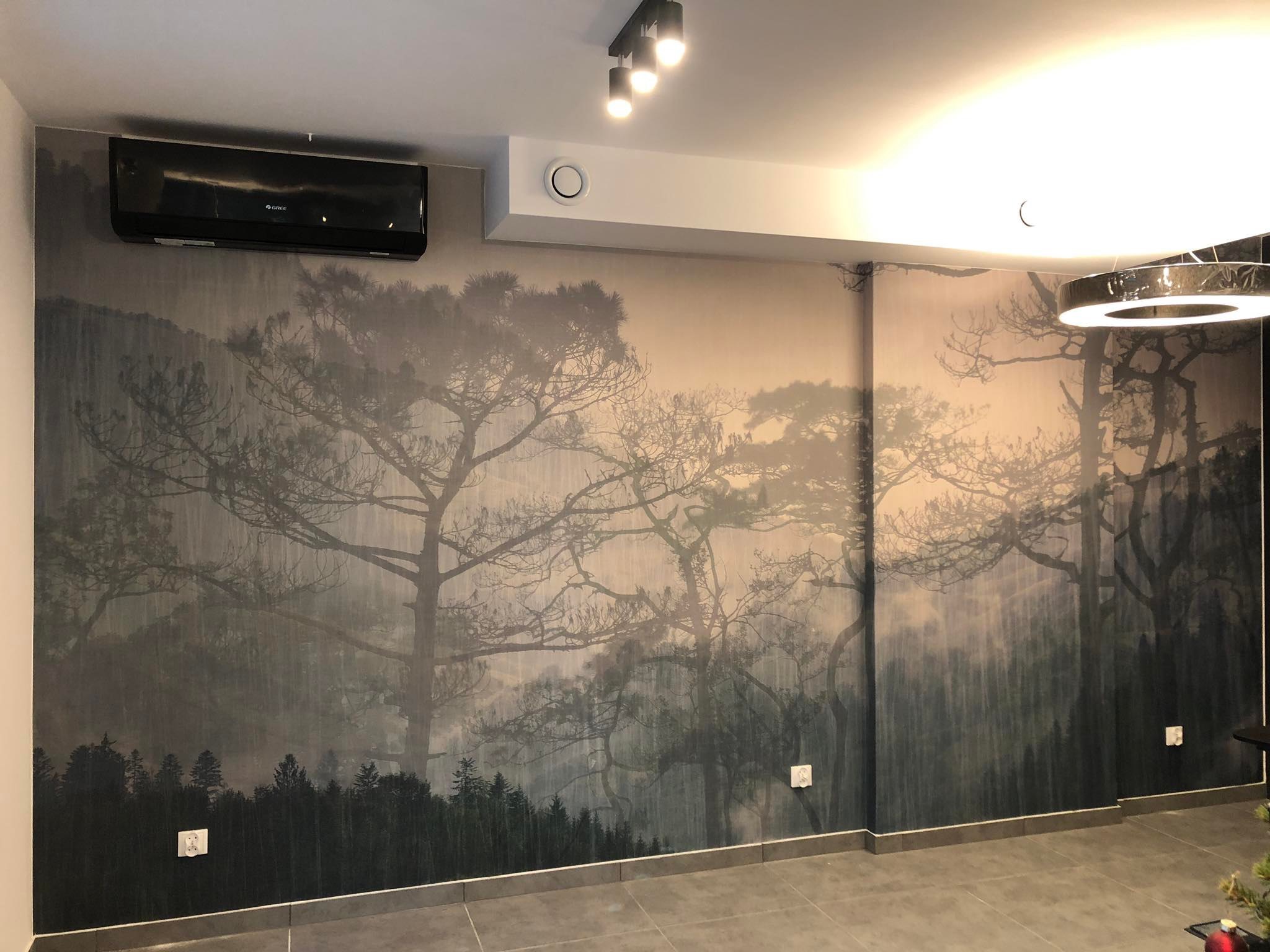 Pokój po tapetowaniu z fototapetą leśną. Na ścianie zamontowany klimatyzator. Szare płytki podłogowe i nowoczesne oświetlenie sufitowe.