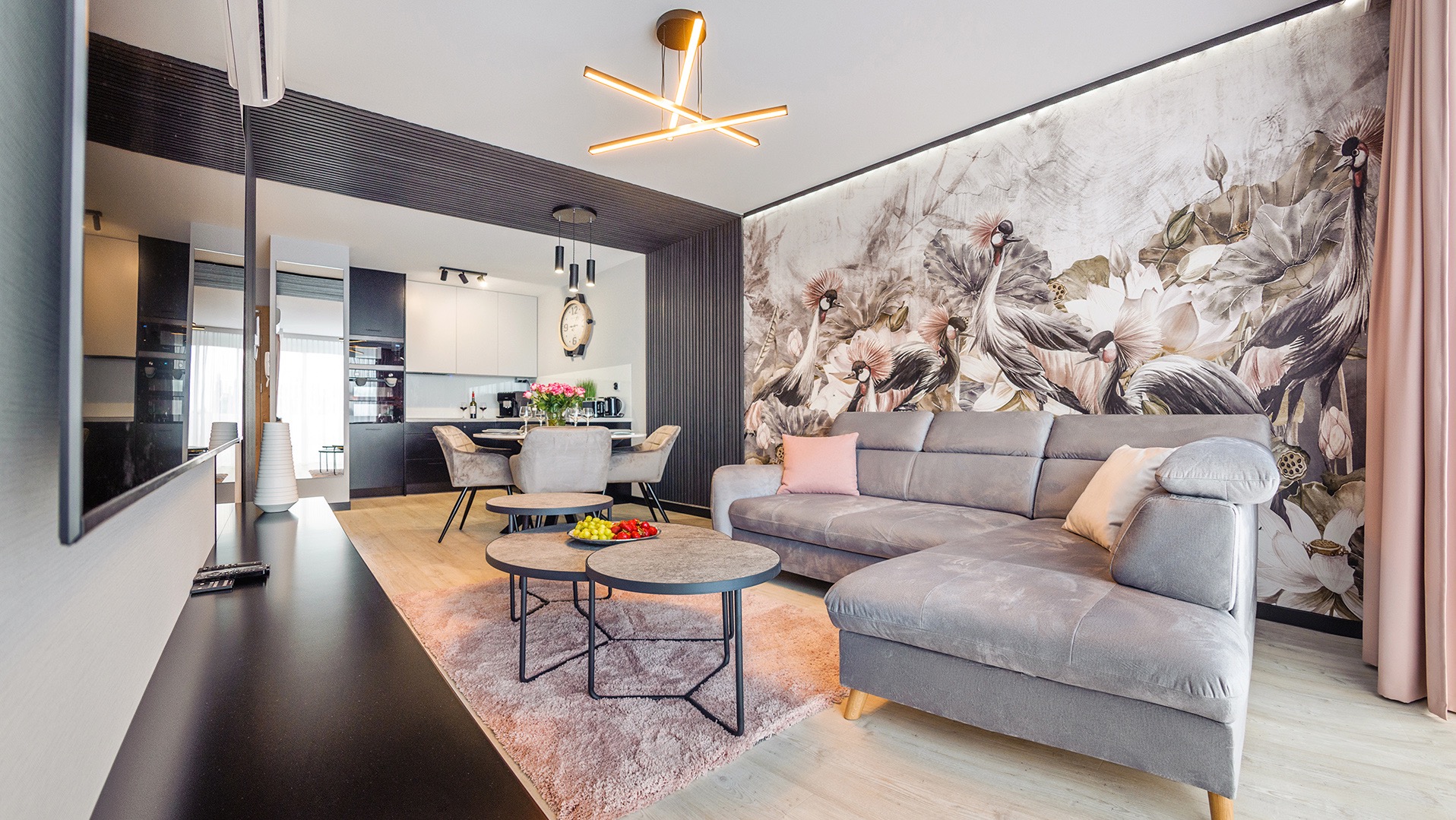 Stylowy salon z tapetą w kwiaty i żurawie, szara sofa narożna, stoliki kawowe i drewniana podłoga. Widok na jadalnię i kuchnię.