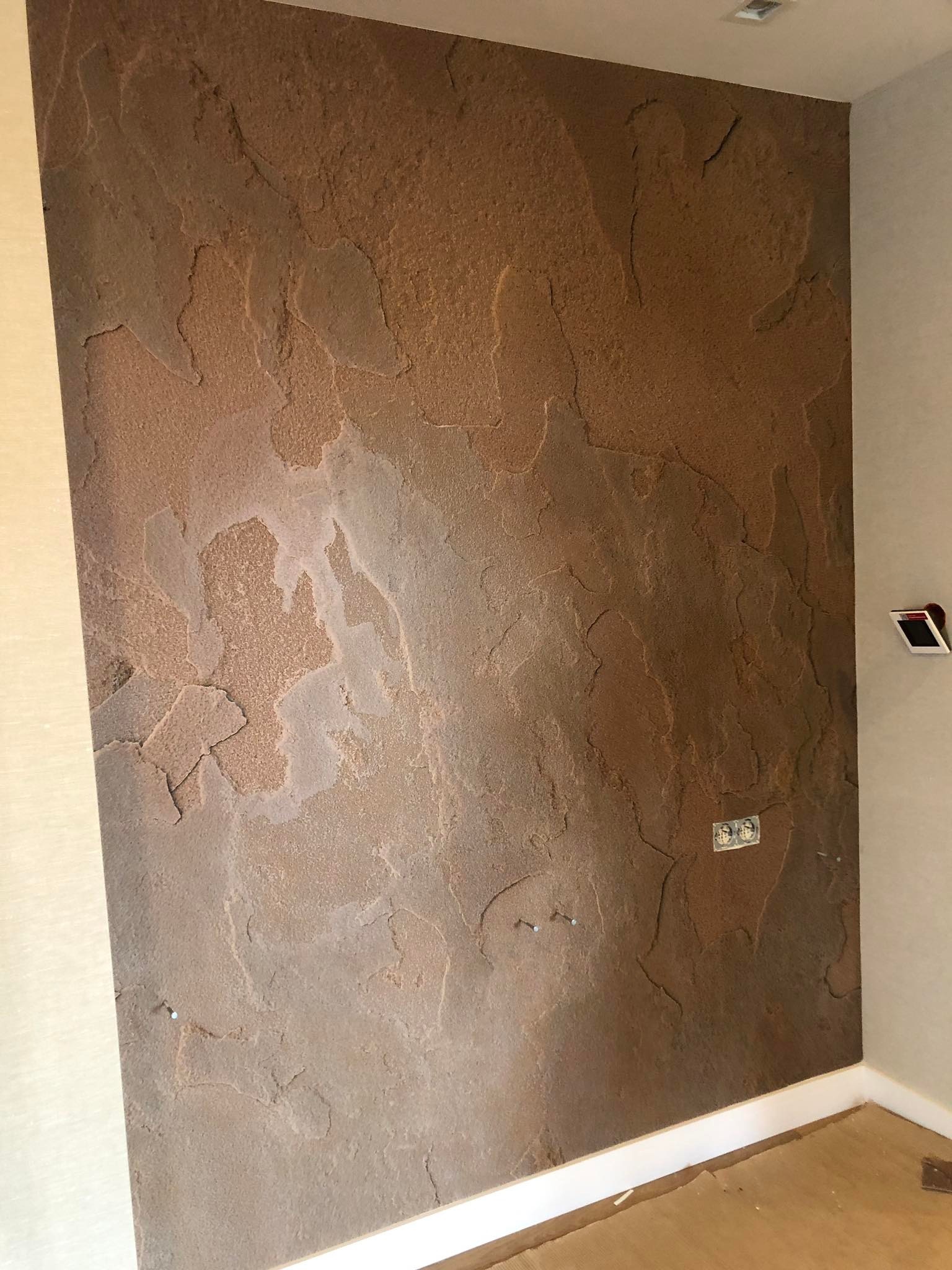 Dekoracyjna ściana w odcieniach brązu z wyraźną, nieregularną fakturą przypominającą naturalny kamień. Widoczne gniazdka elektryczne i listwa przypodłogowa.