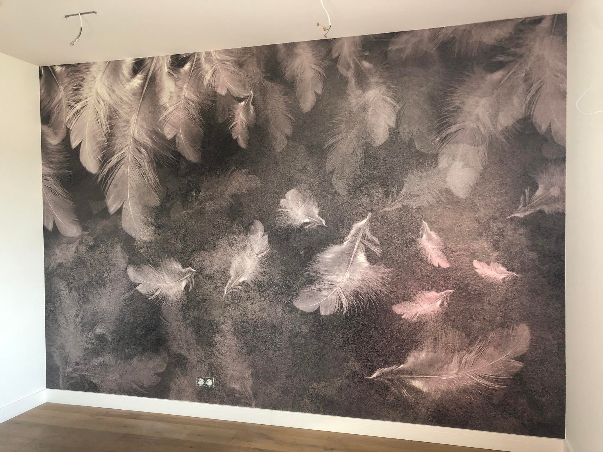 Ściana z fototapetą w motywie piór w odcieniach szarości i różu, widoczne gniazdka elektryczne i fragment drewnianej podłogi, minimalistyczny styl.