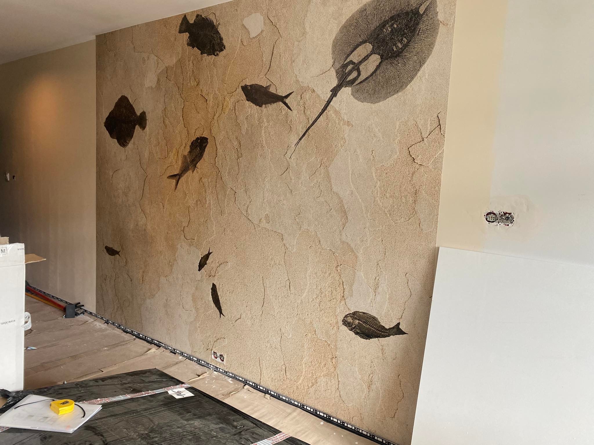Ściana z tapetą imitującą piaskowiec z motywem ryb, w tym płaszczki. Widoczne narzędzia przygotowane do dalszego tapetowania. Realizacja w trakcie prac wykończeniowych.