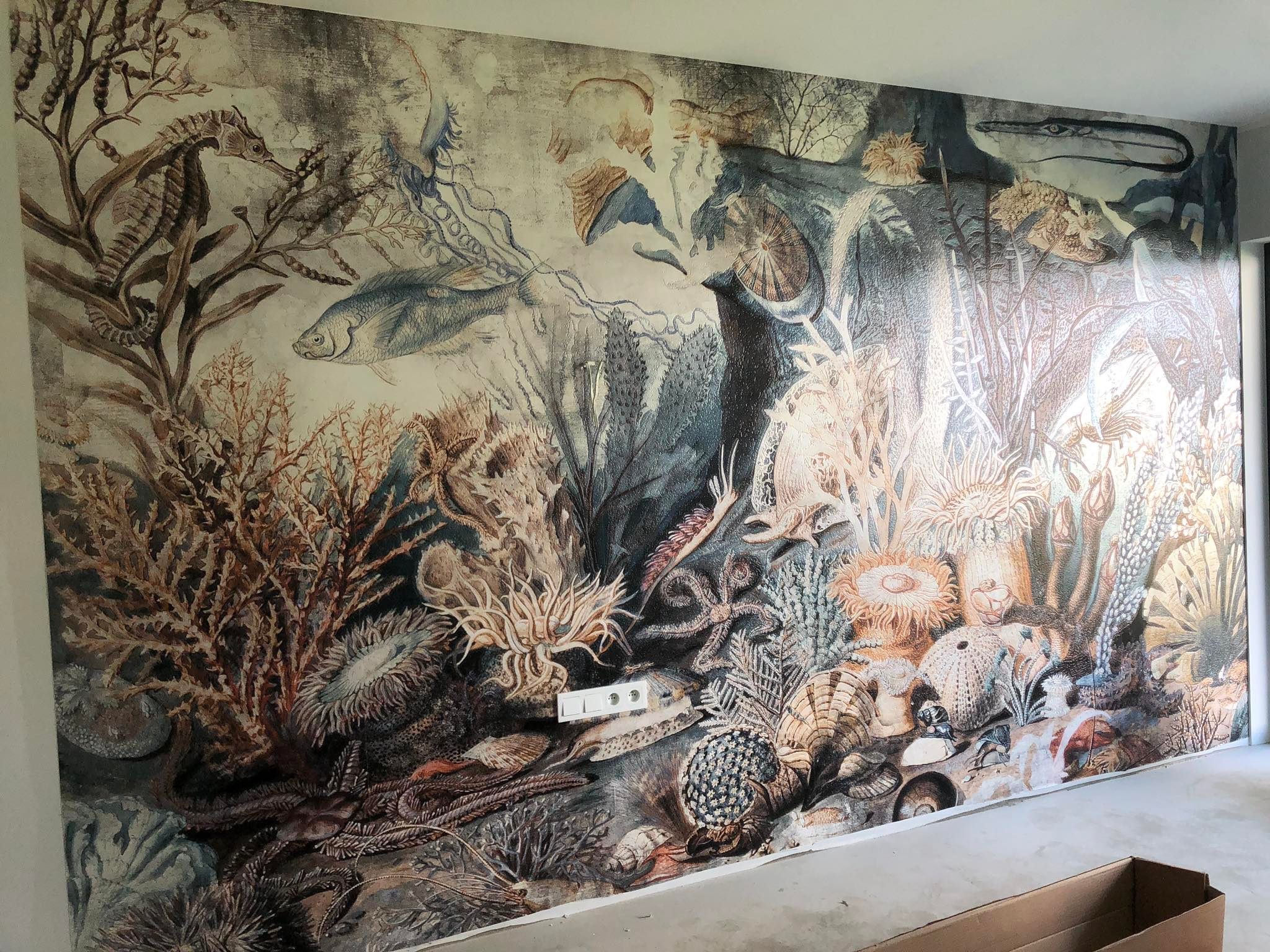 Ściana z tapetą w stylu vintage z motywem podwodnego świata: koniki morskie, ryby, koralowce i muszle. Widoczne gniazdka elektryczne na tapecie.