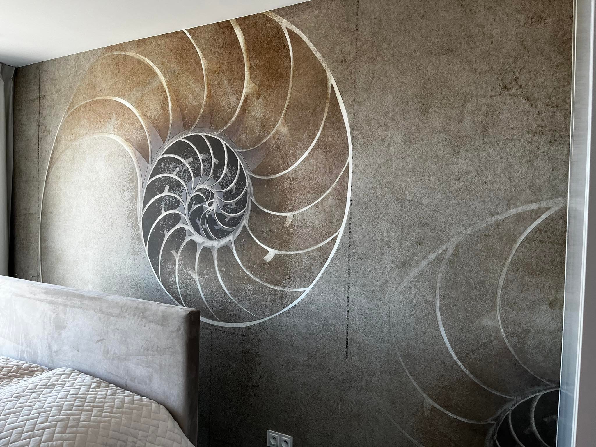 Ściana z tapetą w odcieniach szarości i brązu z motywem muszli nautilusa, widoczny fragment łóżka z szarym zagłówkiem i pościelą w tle.
