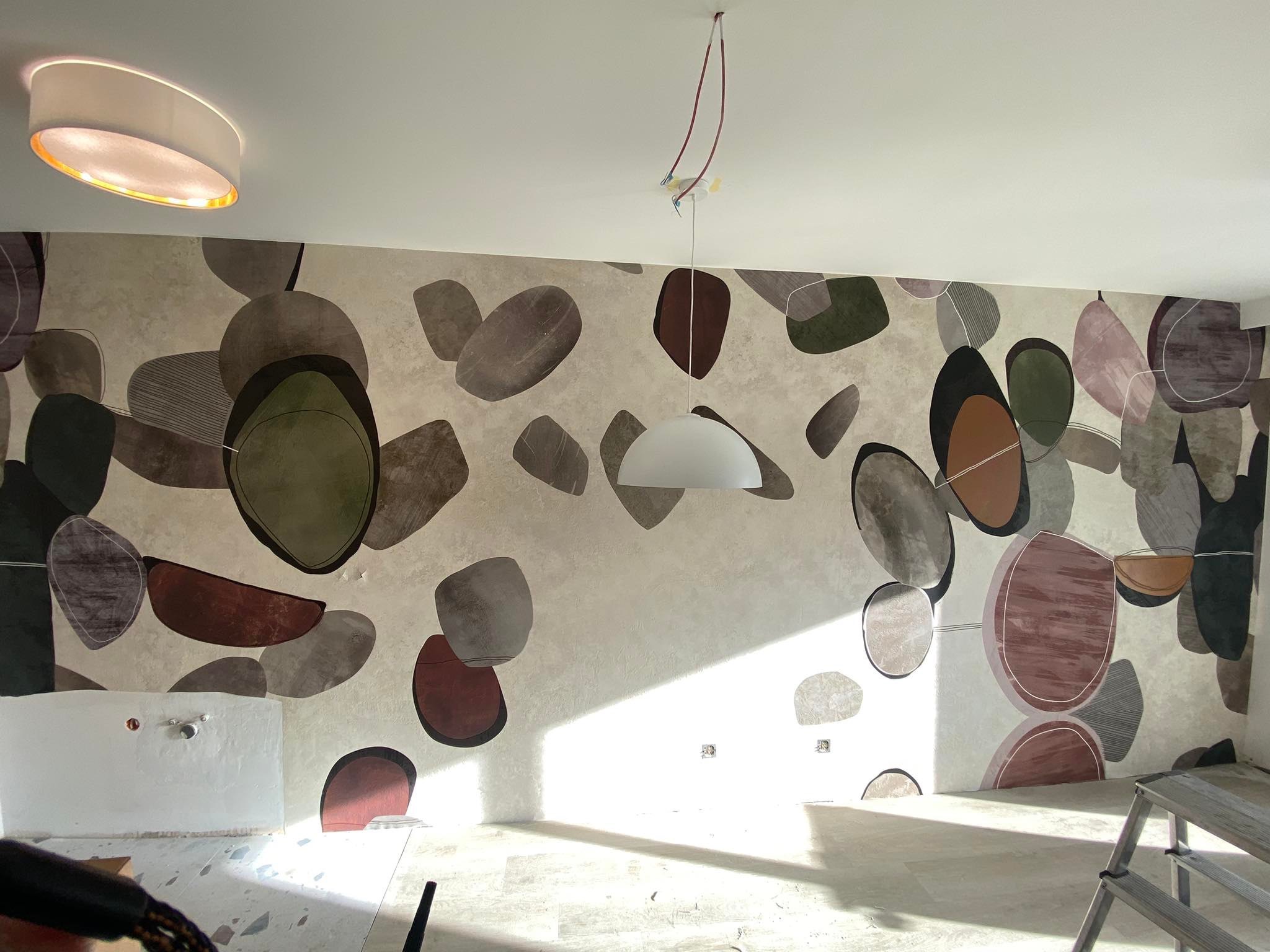 Wykończone wnętrze z tapetą w motywie kamieni w odcieniach brązu i zieleni. Na suficie lampy: okrągła i wisząca. Podłoga jasna, częściowo zakryta. Widoczny fragment drabiny.