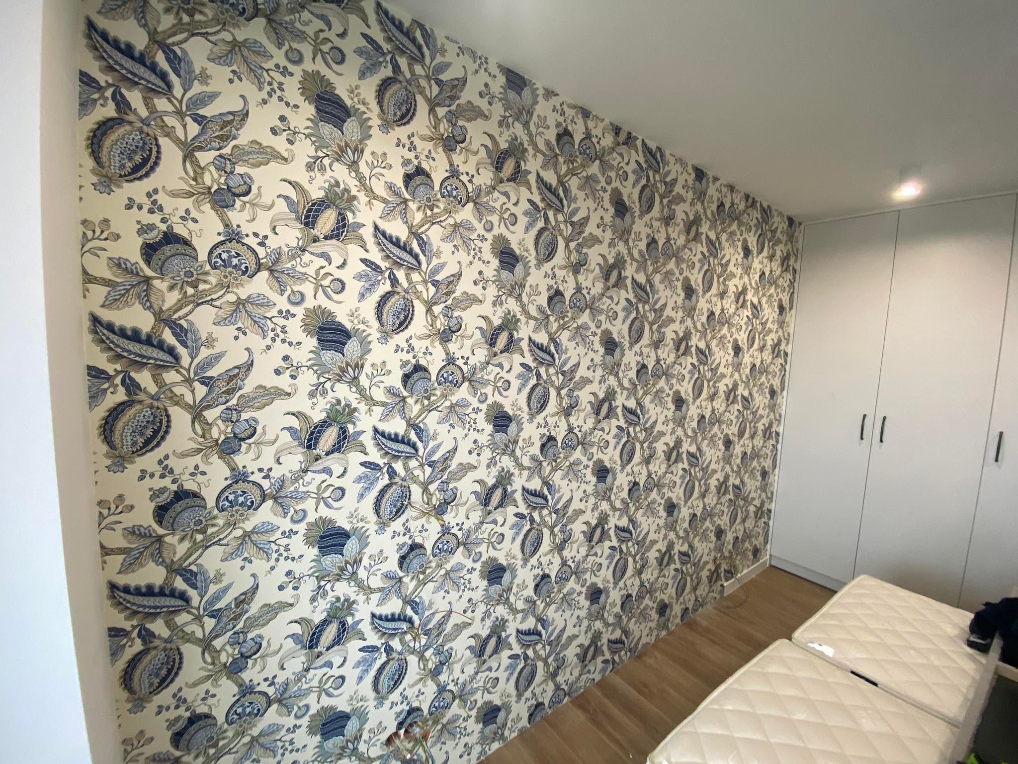 Ściana po tapetowaniu z wzorem roślinnym w odcieniach błękitu i szarości, widoczne fragmenty podłogi i białej szafy wnękowej. Estetyczne wykończenie wnętrza.