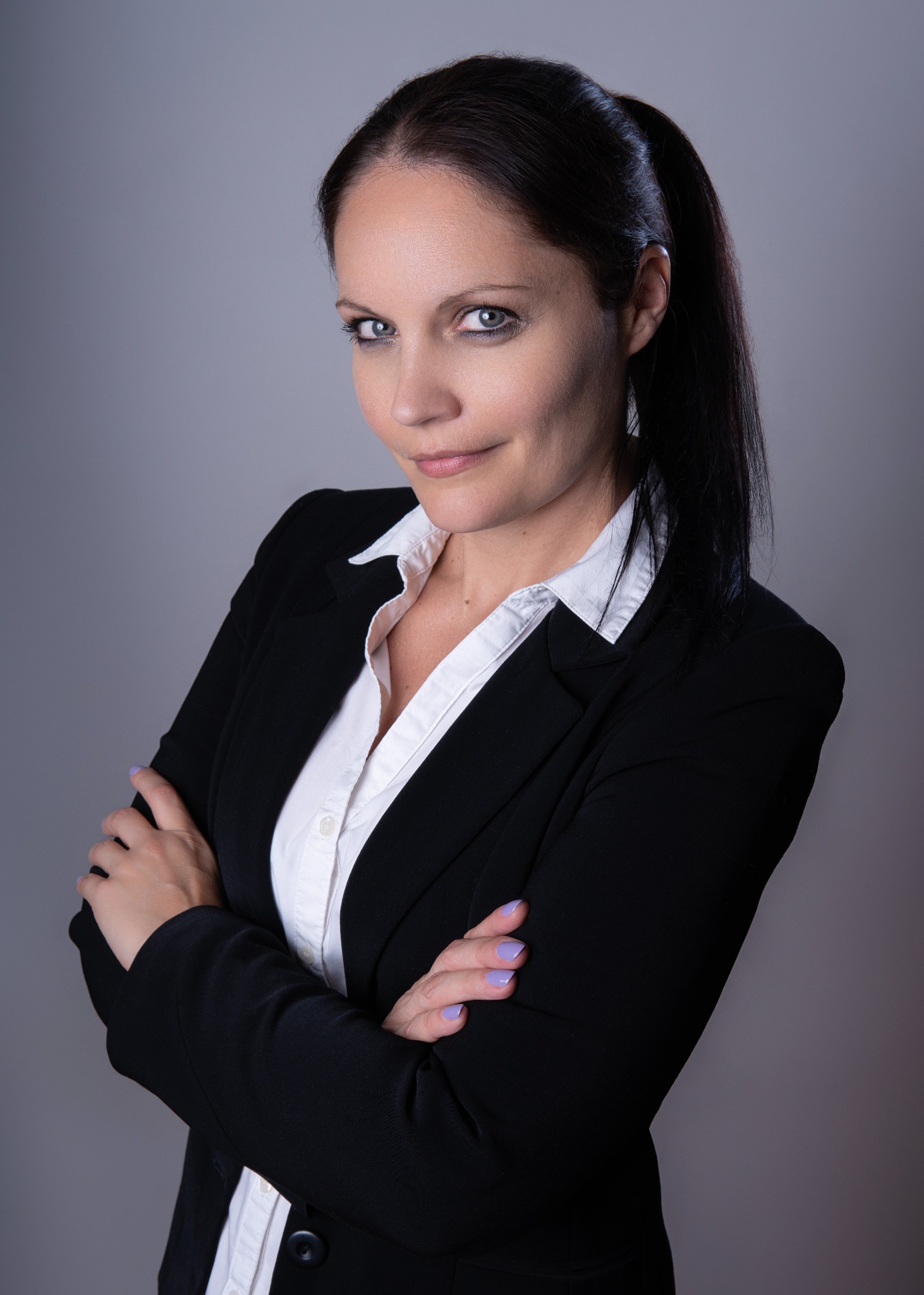 Portret kobiety w czarnym żakiecie i białej koszuli, z założonymi rękami i delikatnym uśmiechem, na jednolitym szarym tle.