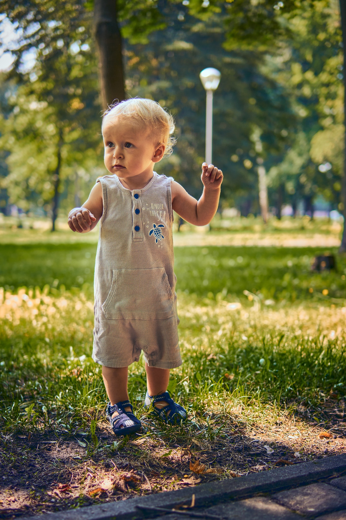 Mały chłopiec w paski kombinezonie z aplikacją żółwia stoi na trawie w parku, w tle widoczna lampa parkowa i drzewa.