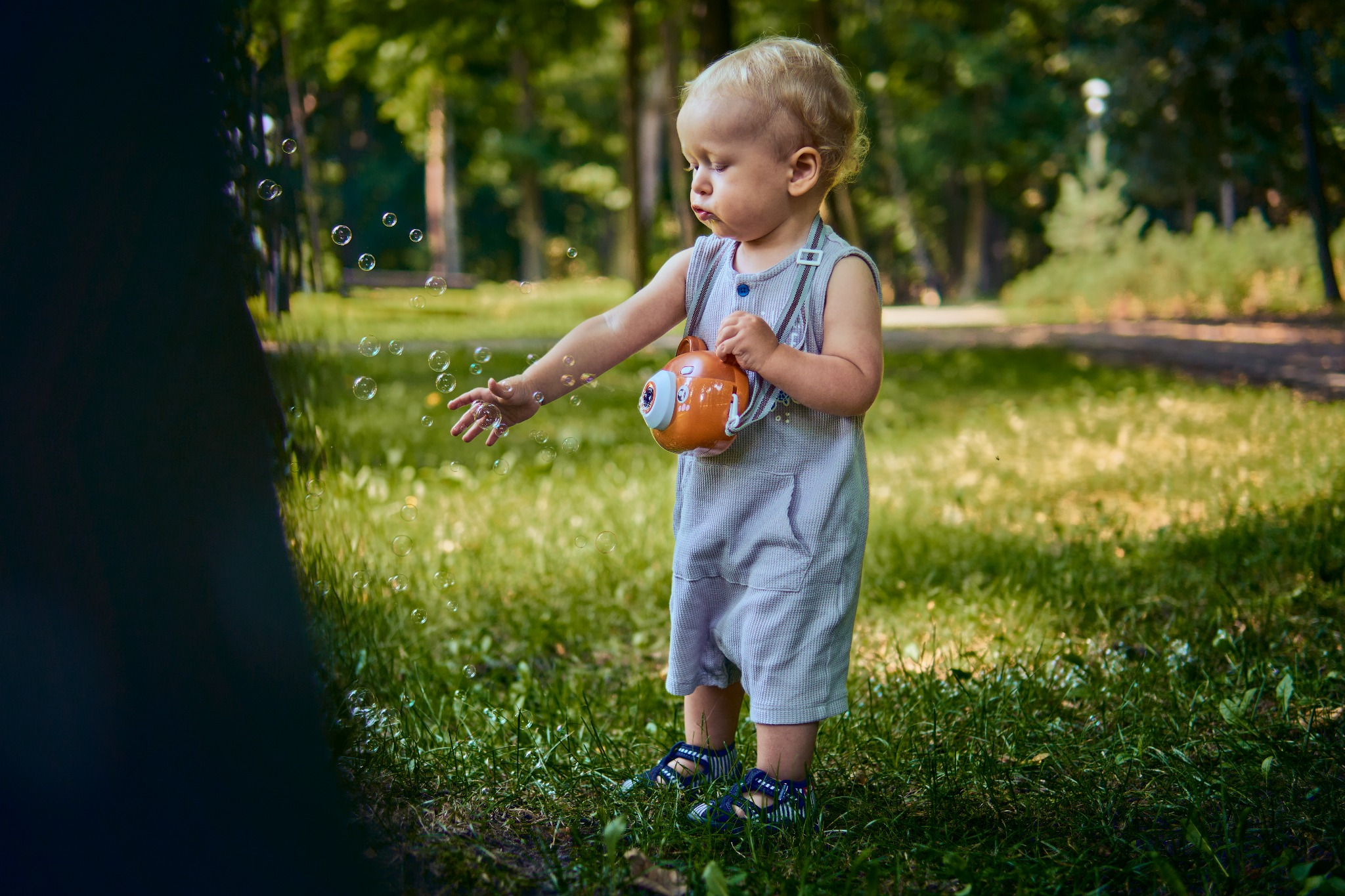Mały blond chłopiec w szarym kombinezonie bawi się w parku, wypuszczając bańki mydlane z pomarańczowej zabawki w kształcie aparatu fotograficznego.