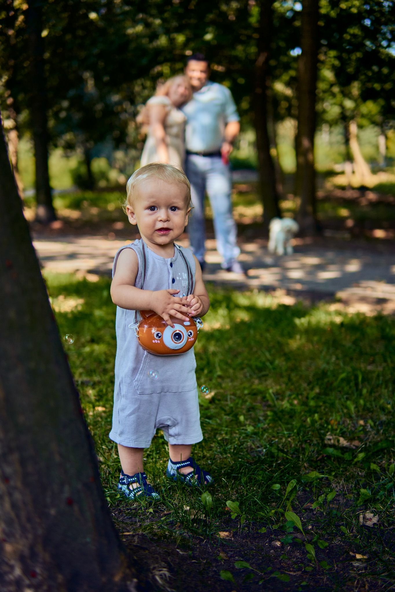 Mały chłopiec w szarym kombinezonie i sandałach, trzymający zabawkę w kształcie aparatu fotograficznego, na trawie z rodzicami w tle, w otoczeniu drzew i puszczanych baniek mydlanych.