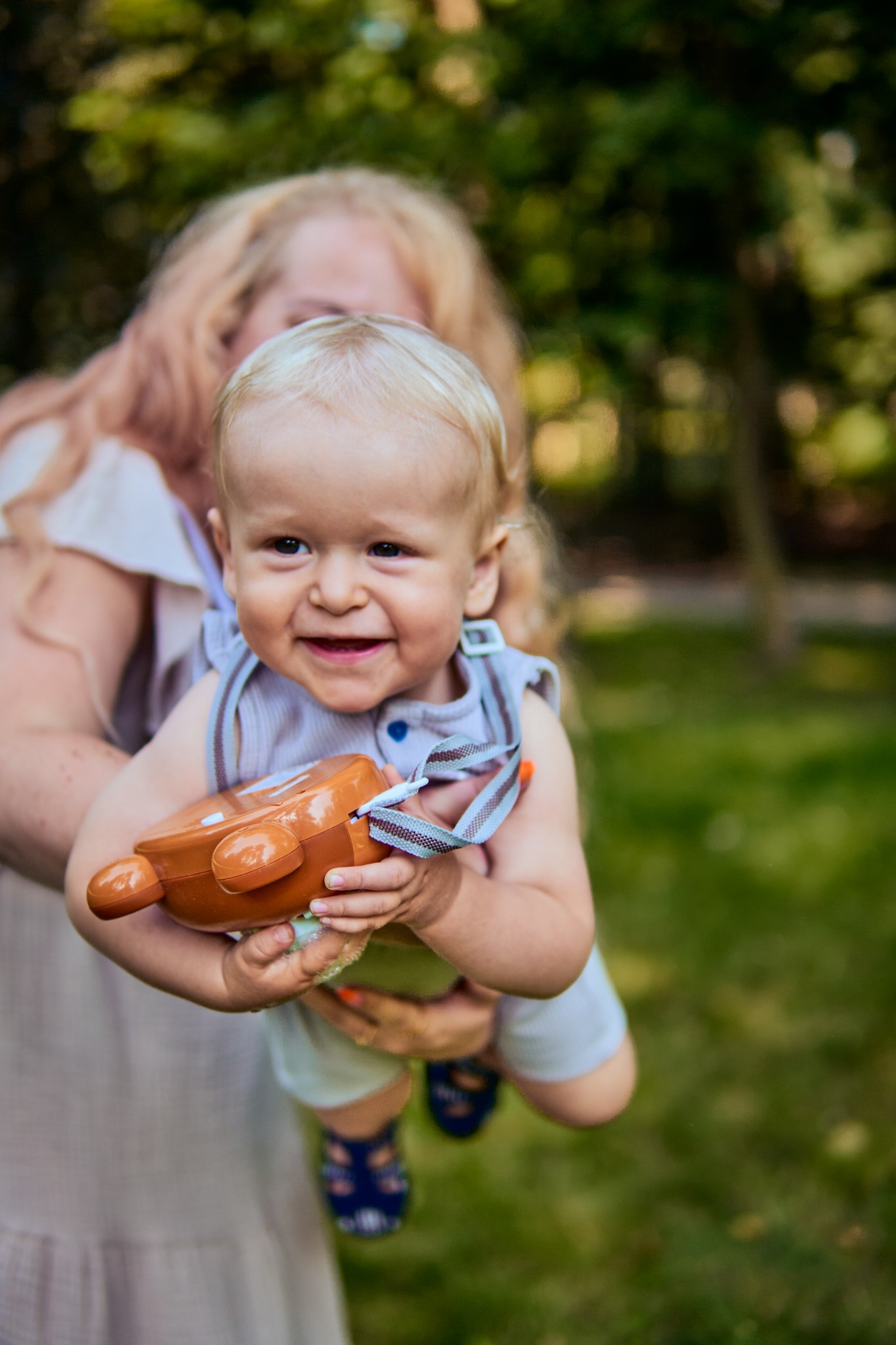 Uśmiechnięty blondwłosy chłopiec trzymany przez dorosłą osobę na tle zieleni, chłopiec ma na sobie letnie ubranie i trzyma brązową zabawkę.