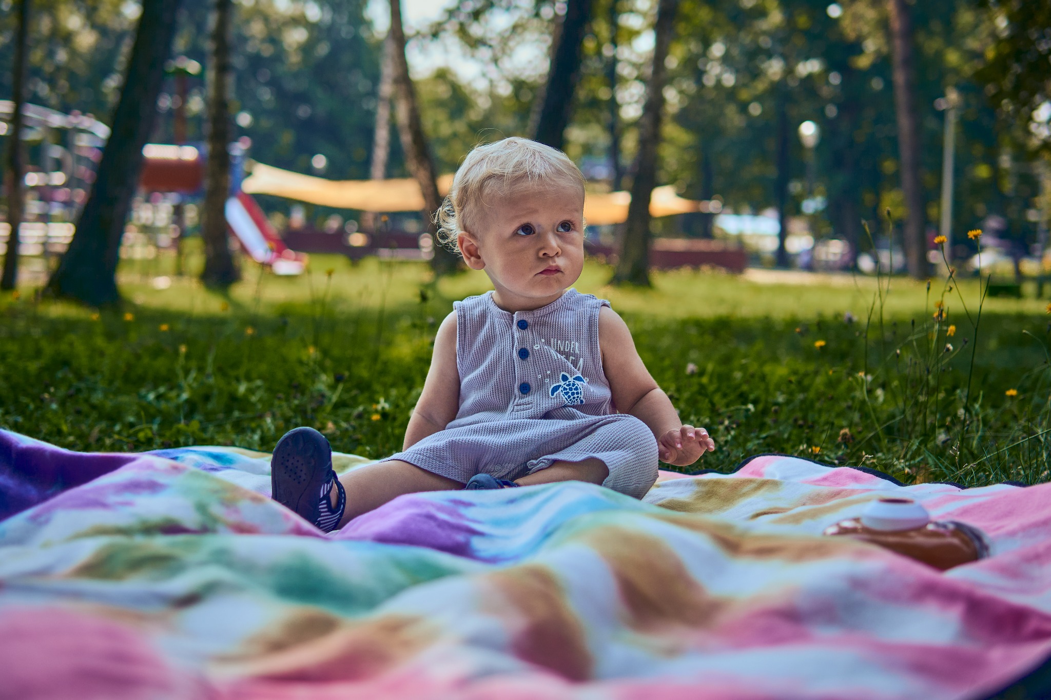 Mały chłopiec w paski siedzi na kolorowym kocu piknikowym w parku, w tle plac zabaw i drzewa.
