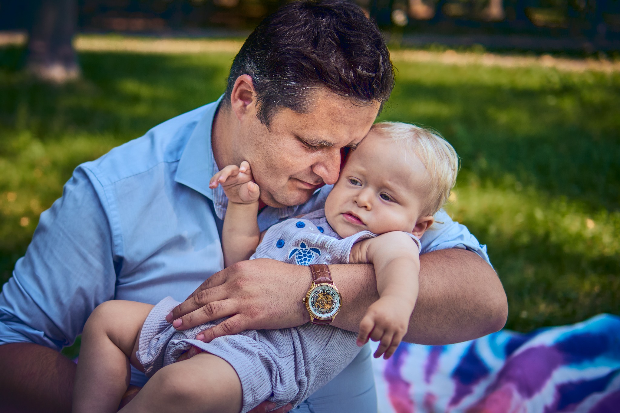 Ojciec w błękitnej koszuli trzyma na rękach małego synka z blond włosami, oboje patrzą w dół, w tle zielona trawa i rozmyte drzewa.